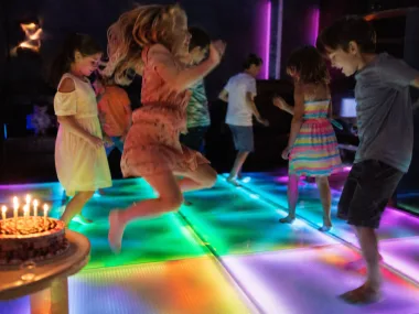 Kinder springen auf der leuchtenden Spielfläche, Geburtstagstorte daneben (KI-Bild)